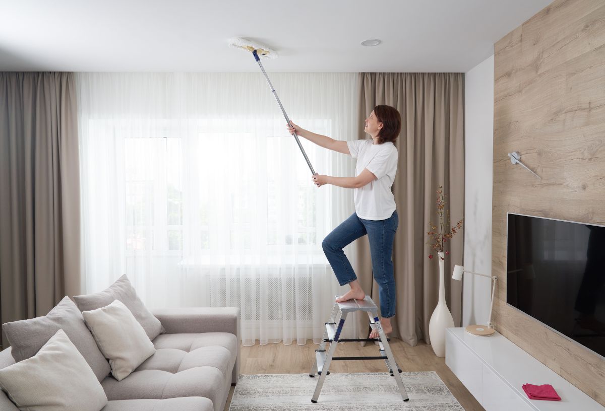 Nő nappali szobában szobalétrán állva seprűvel tisztítja a plafont