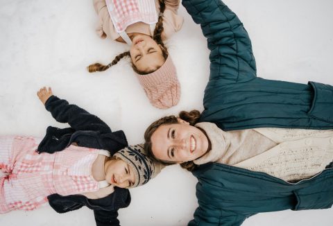 Anyuka kék kabátban mosolyogva fekszik a hátán a hóban két lányával, akik sapkát, kabátot viselnek