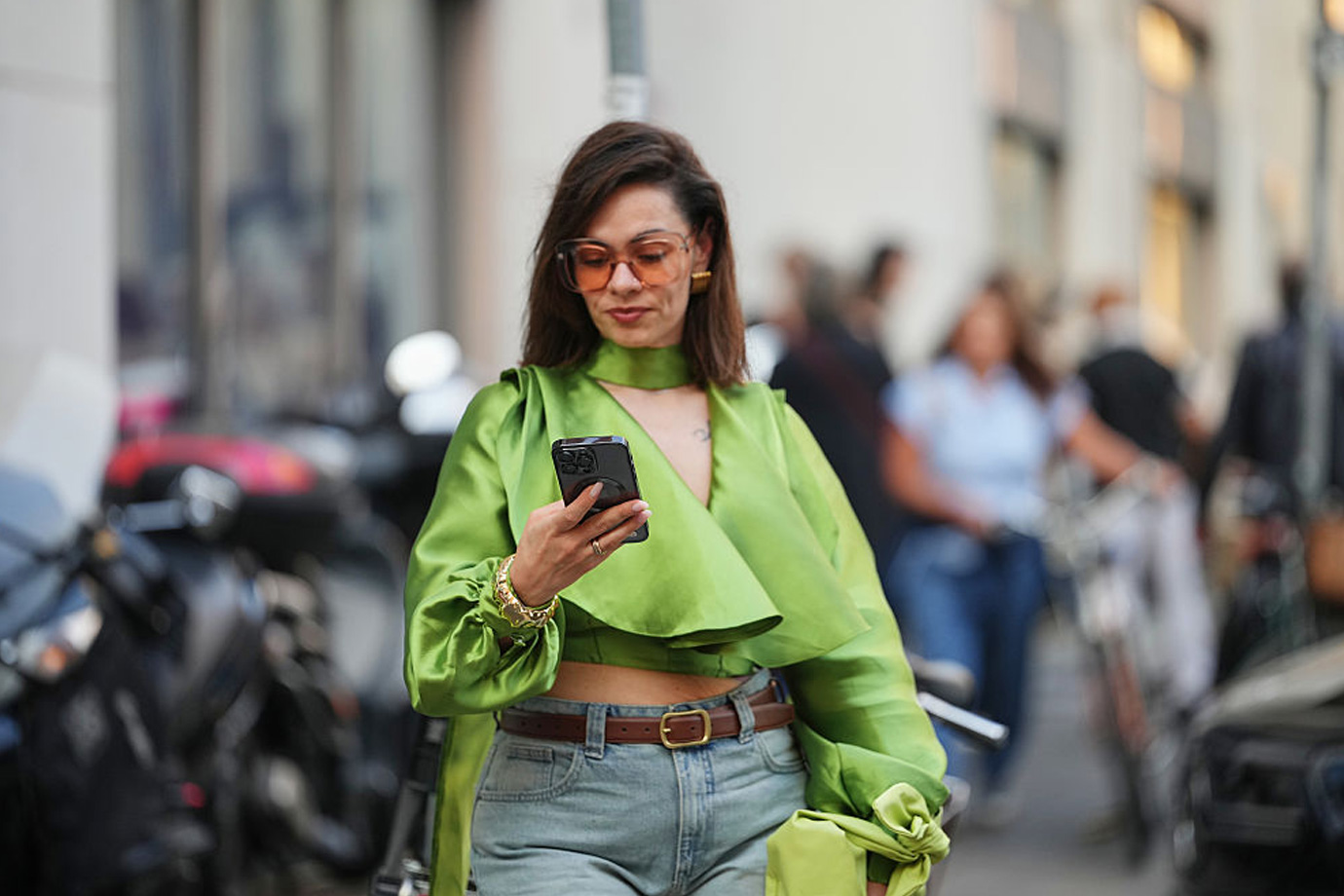 Egy sötétbarna hajú nő, aki túlméretezett, borostyánszínű napszemüveget visel, clutch táskát, valamint sárgászöld szatén blúzt, aminek hosszú, bő ujjai vannak, és bő szárú farmert, egy mobilt tart a kezében.
