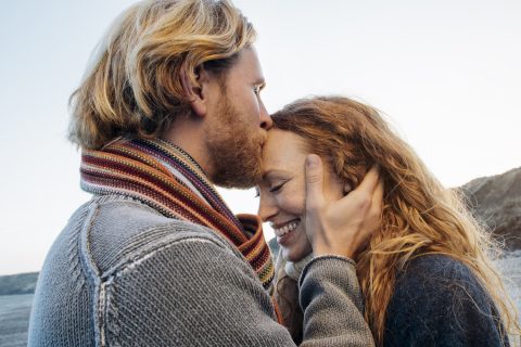 Fiatal pár sétál a tengerparton, a férfi gyengéden tartja a nőt, miközben megcsókolja a homlokát
