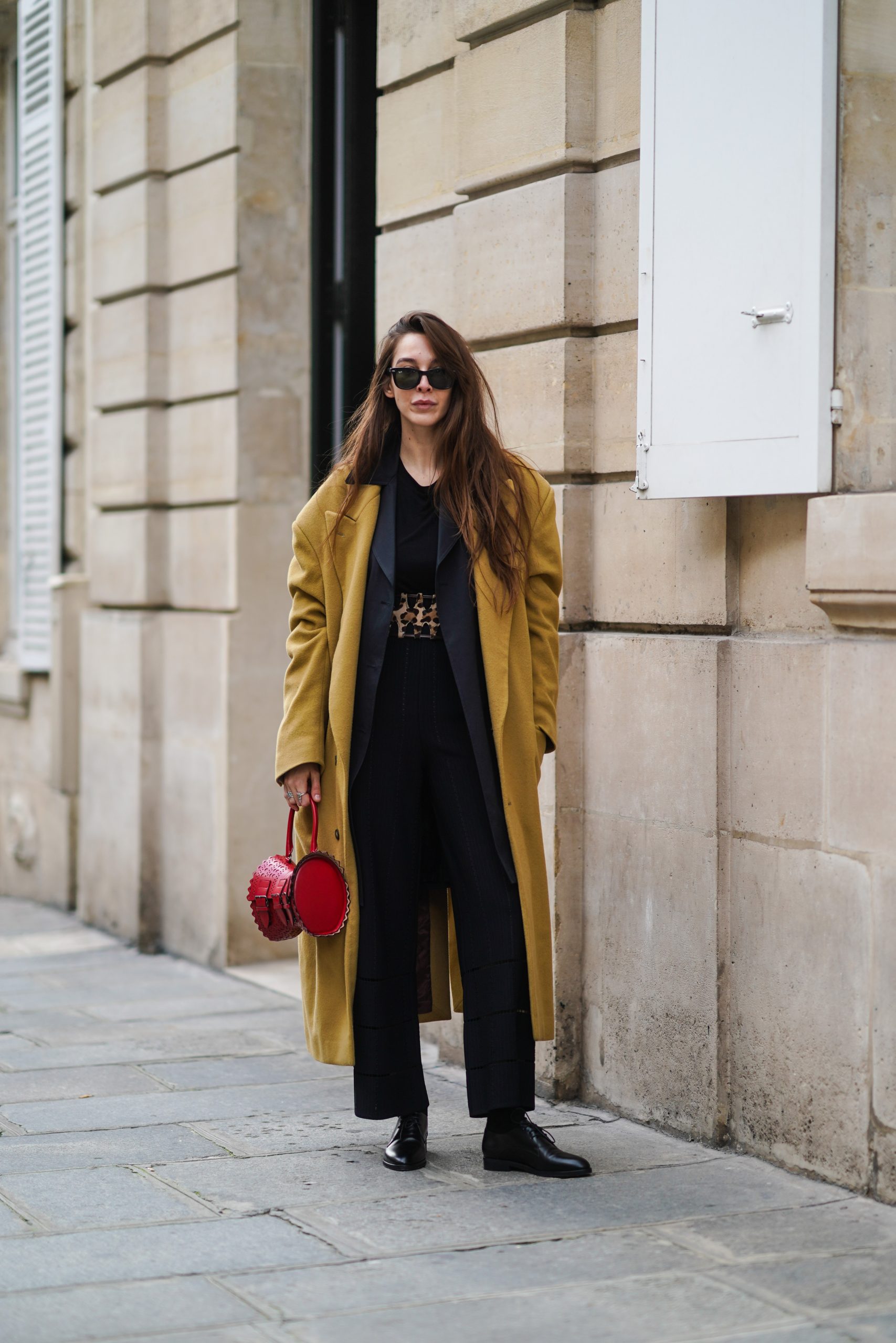 Estelle Chemouny Ray Ban napszemüveget, barna/khaki színű, túlméretezett gyapjú kabátot, piros Alaia bőr táskát, fekete felsőt, sötét szürke, túlméretezett blézert, leopárdmintás széles övet, fekete öltönynadrágot és fekete bőrcipőt visel az Alaïa bemutatóján, a 2021-es párizsi divathéten