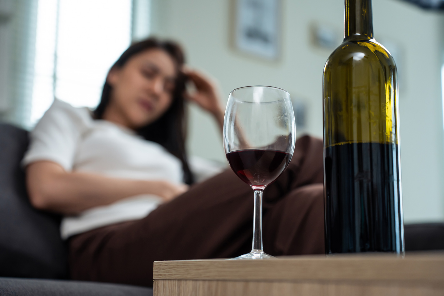 Egy szomorú nő a kanapén ül, előtte egy pohár borral és egy borosüveg az asztalon.