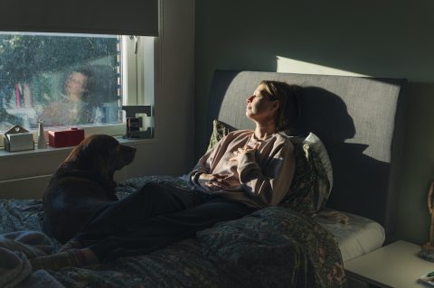 Idősebb nő meditál a reggeli fényben a hálószobájában, miközben házi kedvence, a kutyája figyeli őt az ágyban.