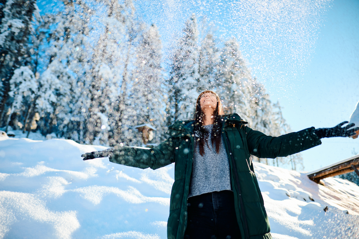 Egy fiatal nő örömmel dobálja a havat a levegőbe, miközben élvezi a téli erdő szépségét, átadva magát a karácsony ünnepi hangulatának és a hóesés varázsának.