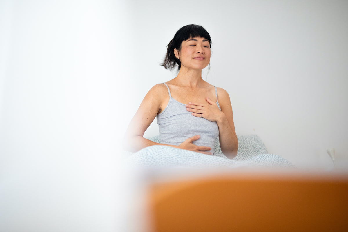 Egy nő az ágyban ülve a légzésére összpontosít, miközben a kezét a mellkasára és a hasára helyezi.