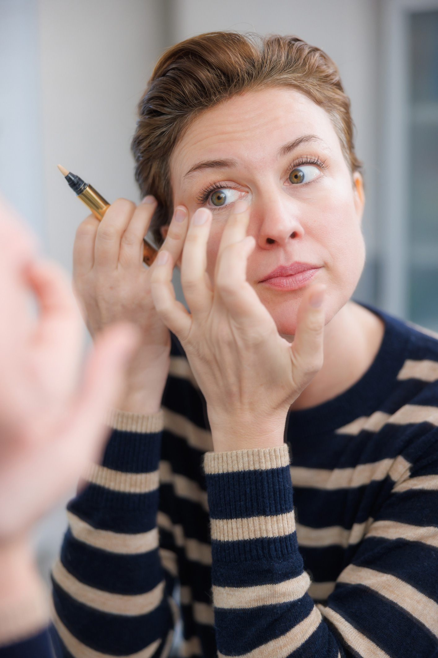 Egy középkorú nő, aki korrektort alkalmaz a szem alatti területre, és sminkel a fürdőszobai tükör előtt