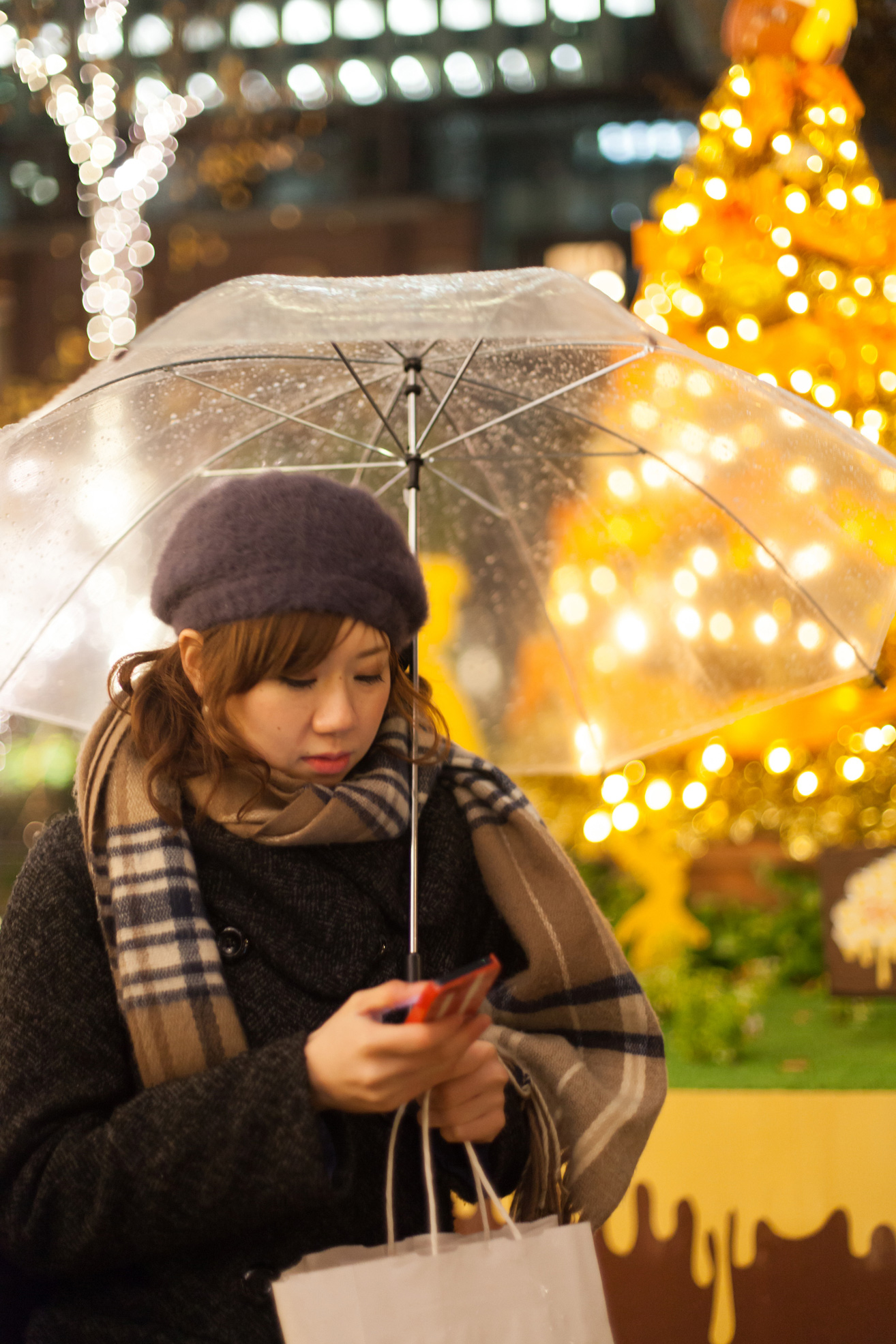 Fiatal nő mobillal és esernyővel a kezében vár az esőben, mögötte feldíszített karácsonyfa
