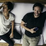 pár a kanapén: a nő a fejét fogja, a férfi fülhallgatón zenét hallgat és nagyon élvezi