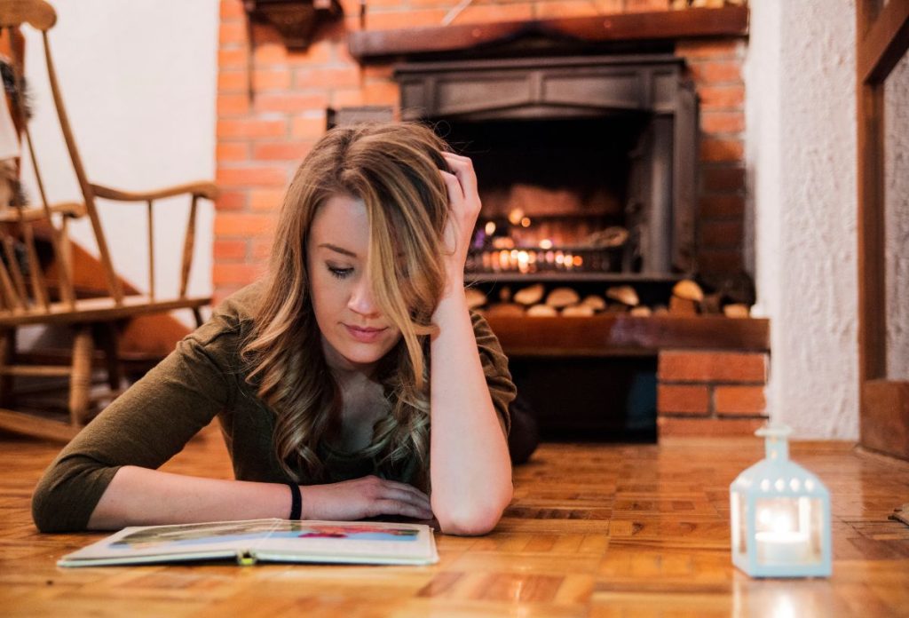 Szép fiatal nő könyvet olvas hangulatos téli szobában kandalló előtt hasalva