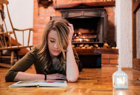 Szép fiatal nő könyvet olvas hangulatos téli szobában kandalló előtt hasalva