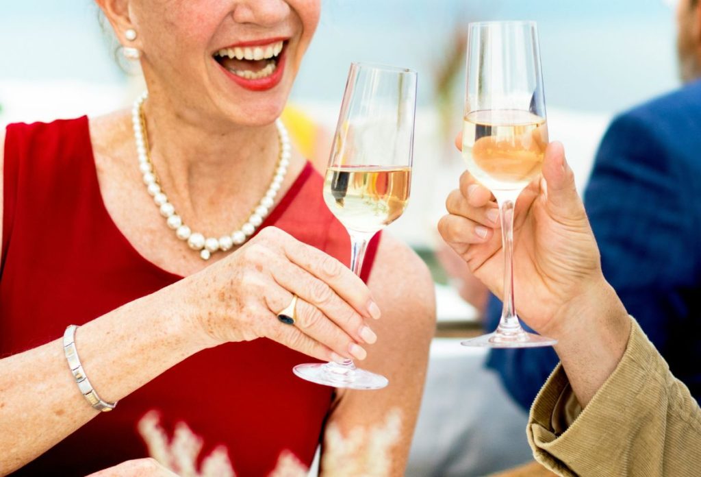 Idős nő piros felsőben, fehér gyöngynyakláncban, piros rúzzsal pezsgővel koccint férfival