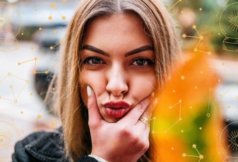 Nő csücsörítve fogja az arcát őszi hangulatú háttér előtt, asztrológiai jelképek a háttérben