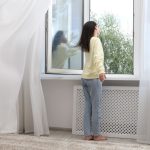 Barna hajú nő farmerben és fehér pulóverben a nyitott ablaknál áll, hosszú fehér függönyt fúj a beáramló szél