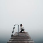 Móló végén a ködbe tekint egy sapkás, földön ülő nő