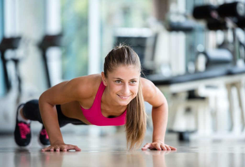 Fiatal, mosolygós nő rózsaszín sportfelsőben fekvőtámaszt végez az edzőteremben
