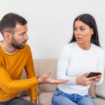 Férfi narancssárga pólóban és nő kék farmerben és fehér pólóban, kanapén ülnek, és vitatkoznak