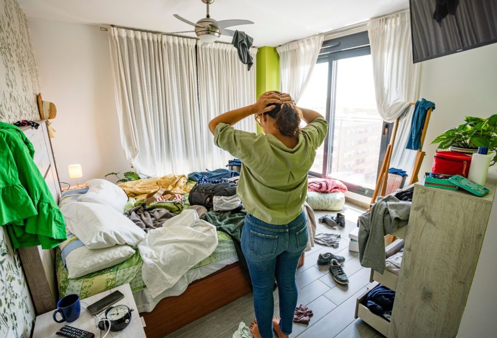 Nő farmerben és zöld ingben rendetlen szobában áll tehetetlenül