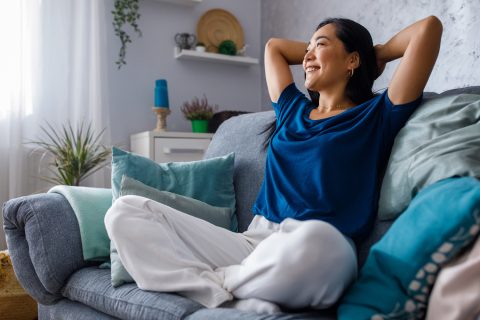 Egy gyönyörű, boldog fiatal nő, aki a kényelmes kanapén ül, kezeit a feje mögé emeli, miközben mosolyog és álmodozik