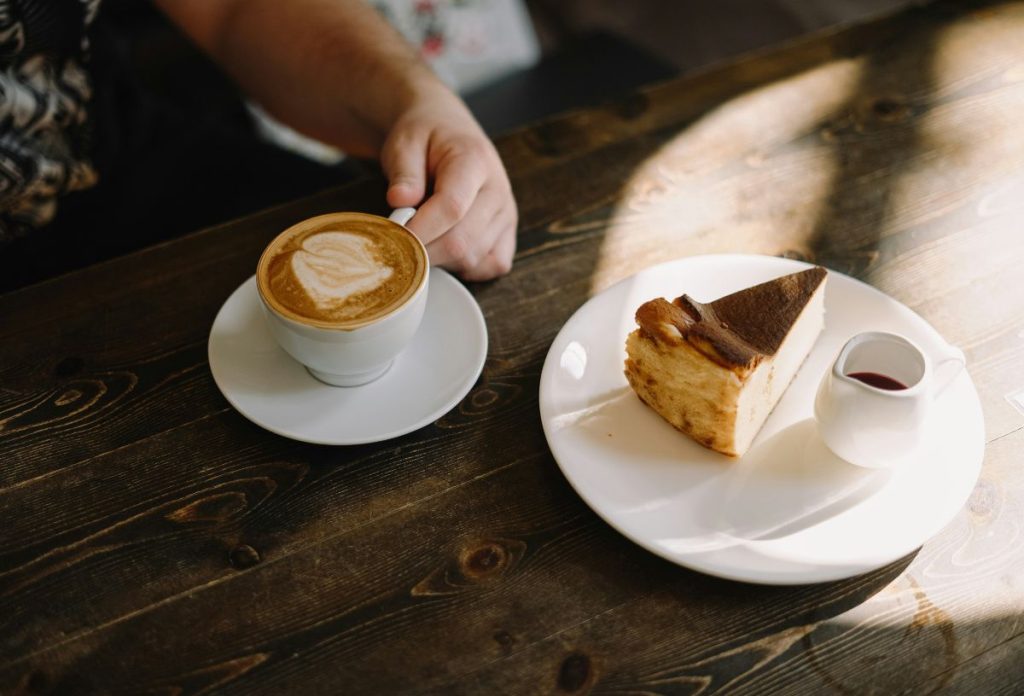 Férfi kávéscsésze fülét fogja, mellette fehér tányéron sajttorta