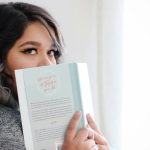 könyvet tartó nő eltakarja a száját a regénnyel és kíváncsian néz