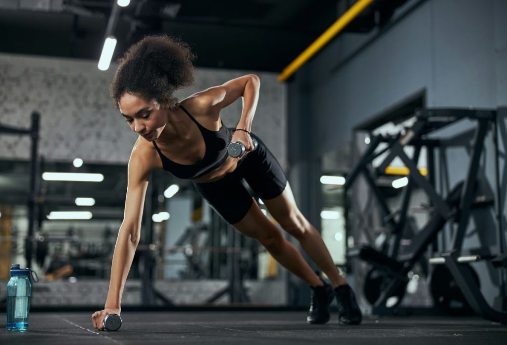 Fekete nő kis súllyal a kezében végez planket evezéssel edzőteremben