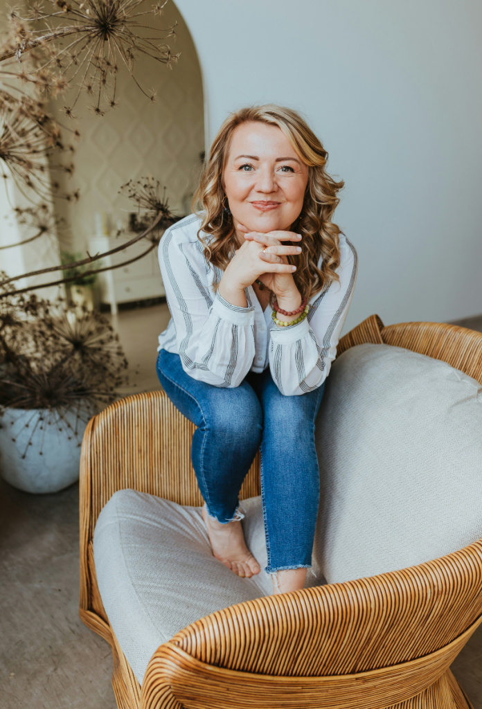 Szép, szőke hajú negyvenes éveiben járó nő farmerben ül fotel karfáján mezítláb