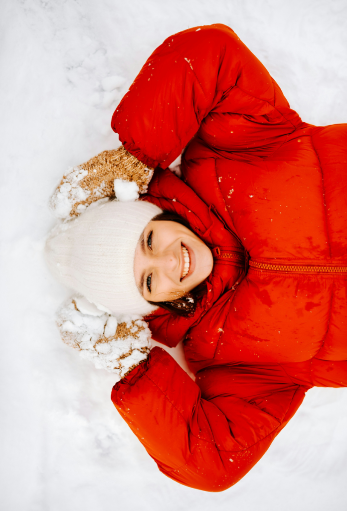 Nő piros télikabátban, fehér sapkában és sárga kötött kesztyűben fekszik a hóban