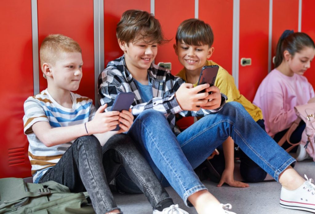 Fiúk a szekrényüknek dőlve mobiltelefonoznak az iskolában a földön ülve