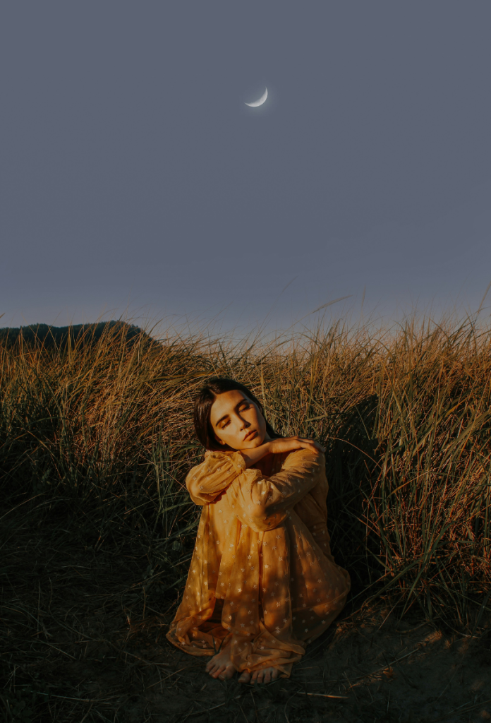 Fiatal, szép nő felhúzott térdekkel ül a fűben, égbolton újhold