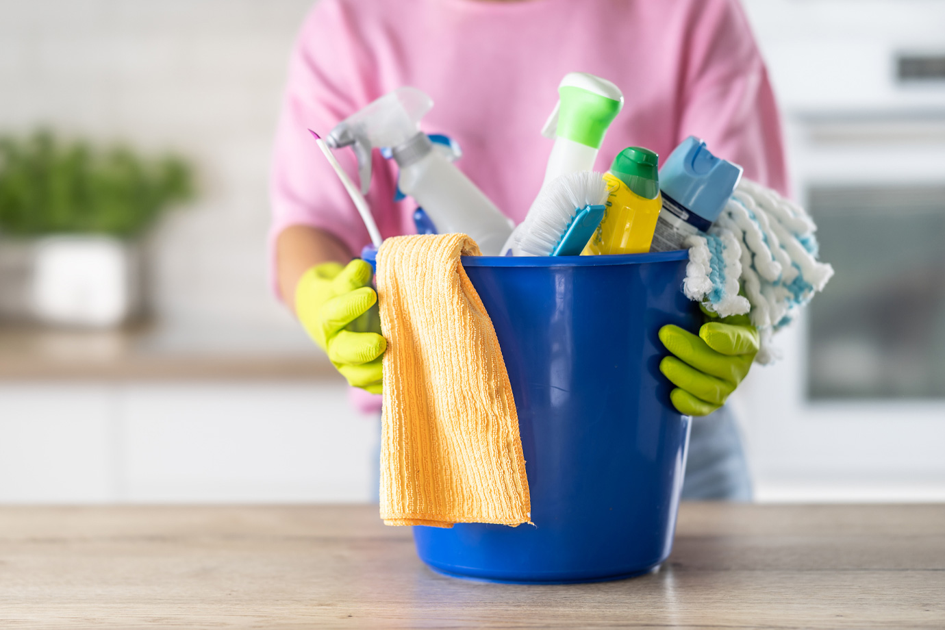 Egy tisztítószerekkel teli vödör, amelyet egy gumikesztyűt viselő nő tart a kezében