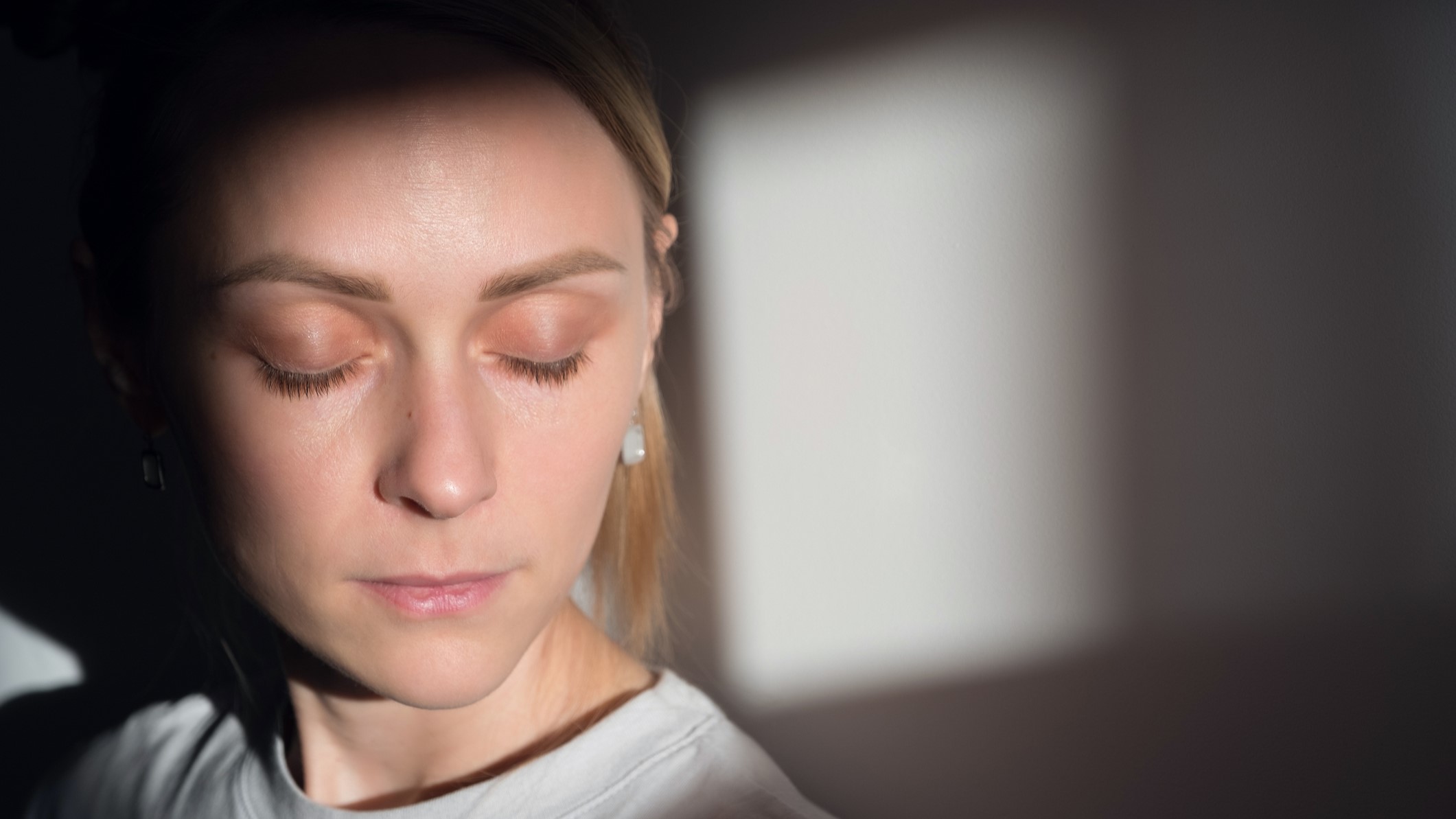 Egy magányos, szomorú, nyugodt lány vagy nő áll egy fehér fal előtt, egy pszichiátriai klinikán vagy otthon, a napfényben, csukott szemmel.