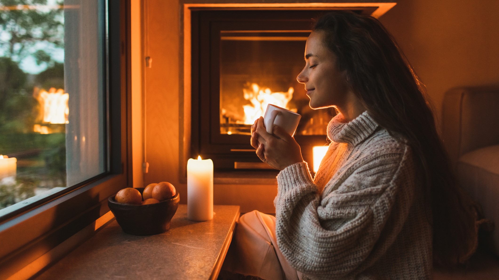 Gyönyörű fiatal hölgy pihen otthon a kandalló mellett. Égő gyertya, tál mandarinnal, karácsonyi hangulat