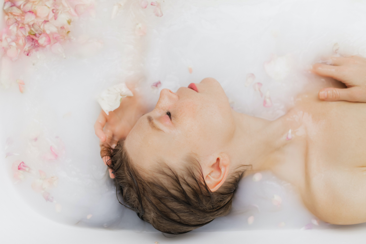 nő kád fehér vízben fekszik, rózsaszín szirmok vannak körülötte