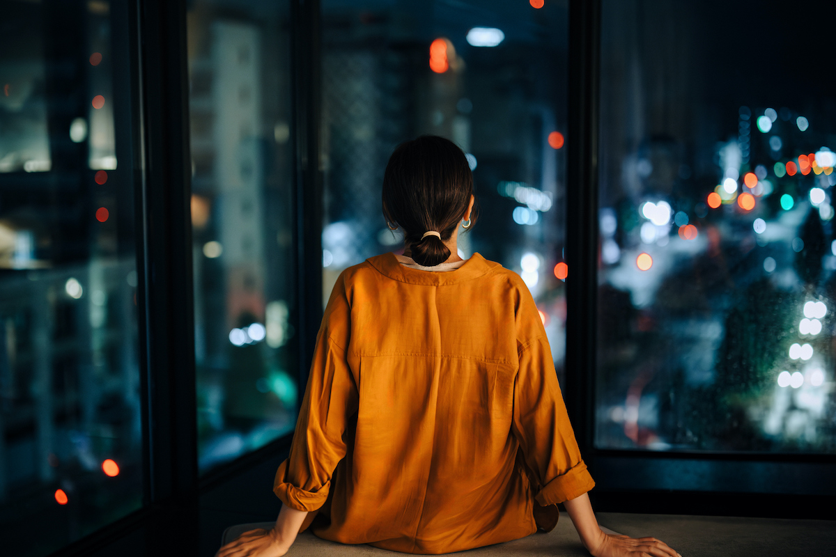 Fiatal nő hátulnézetből, aki otthona meghitt terében pihen: egyedül ül a nappaliban, és az ablakon át a lenyűgözően kivilágított éjszakai város panorámáját nézi.