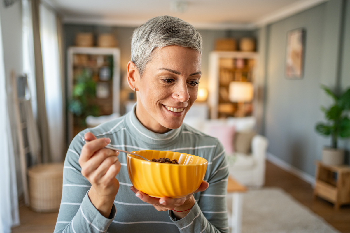 rövid, ősz hajú nő sárga tálból kanalaz magvakat, granolát és mosolyog
