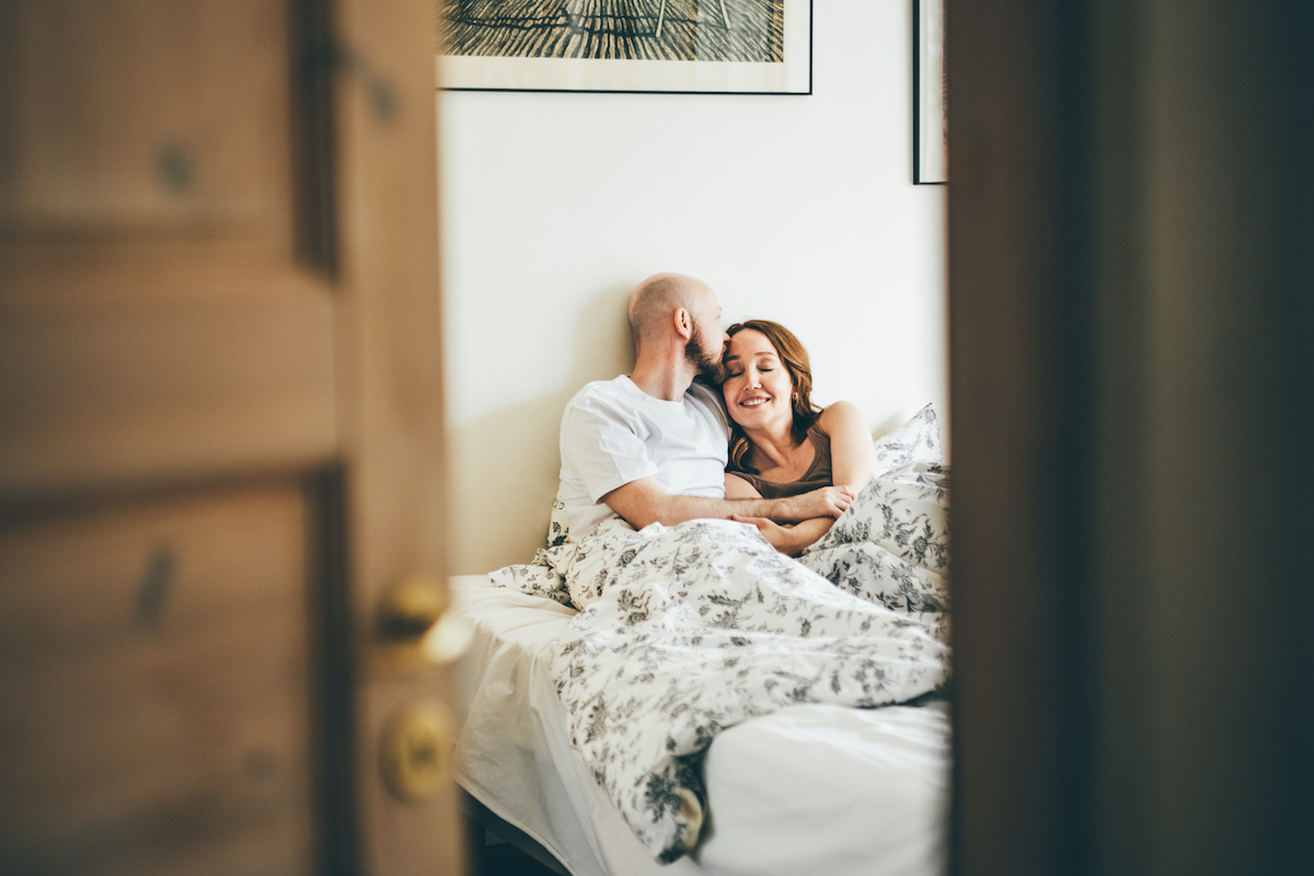 ágyban ül egy pár, a férfi megcsókolja a nő fejét. nyitott ajtón keresztül láthatóak