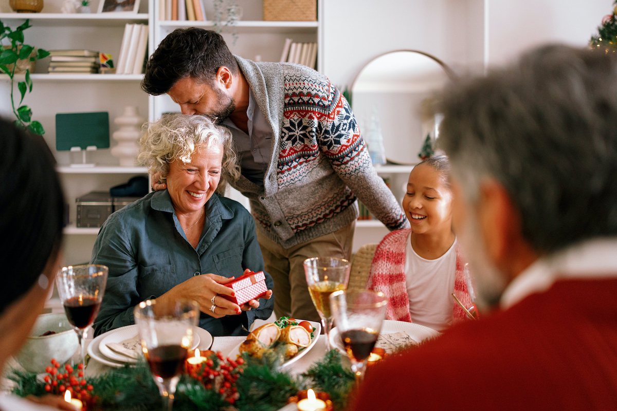 hogyan-mondj-nemet-az-anyosodnak-3-mondat | Marie Claire karácsonyi asztalnál ülve mosolygó fiú megpuszilja az anyját és ajándékot ad