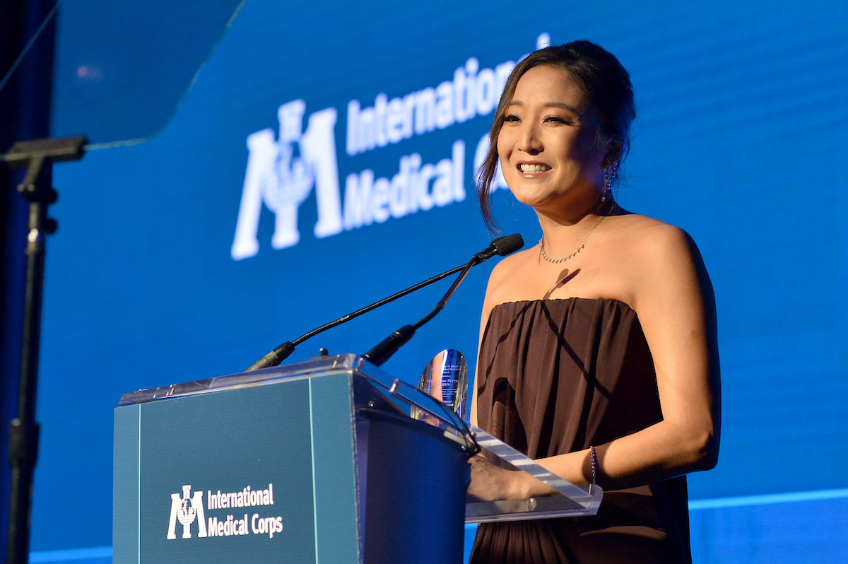 emily-parizsban-ok-voltak-lucas-bravo-szerelmei-az-eletben | Marie Claire 2025. november 13. Ashley Park az International Medical Corps' 2025 Annual Awardson, Beverly Hillsben beszél a színpadon, kék háttér előtt.