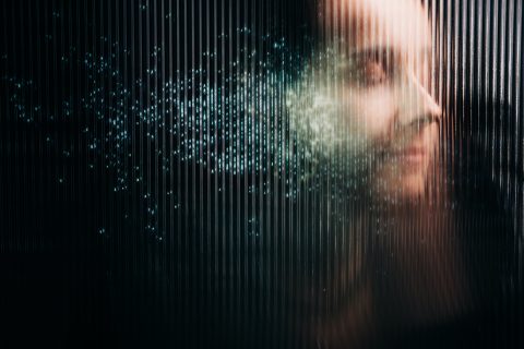 Homályosan kirajzolódó emberi profil látható egy függőlegesen bordázott felület mögött, amely részben fénylő, kék digitális részecskék felhőjévé oszlik. A kép konceptuális és sötét hangulatú, a digitális identitást, az adatvédelmet vagy az emberi tudat töredezését jelképezi a technológia korában.
