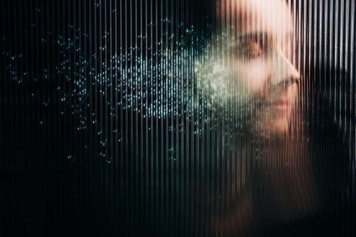 Homályosan kirajzolódó emberi profil látható egy függőlegesen bordázott felület mögött, amely részben fénylő, kék digitális részecskék felhőjévé oszlik. A kép konceptuális és sötét hangulatú, a digitális identitást, az adatvédelmet vagy az emberi tudat töredezését jelképezi a technológia korában.