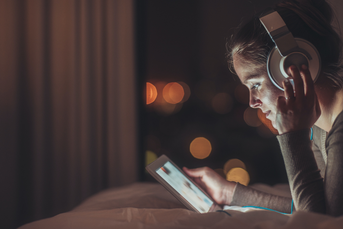nő este az ágyon fekve fülhallgatót hallgat és a telefonon zenét válogat