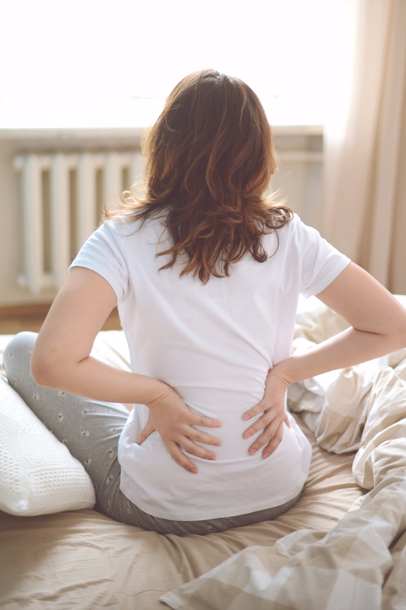 Egy fehér pólós nő reggel a derekát fogja az ágyán