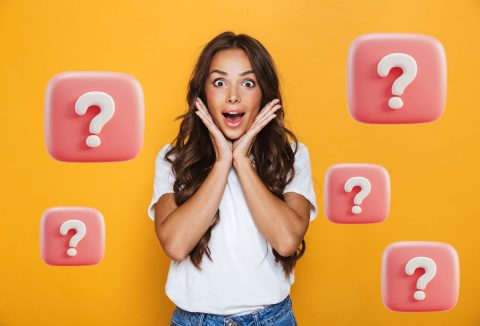 Fiatal, szép nő, hosszú barna hajjal, fehér pólóban, farmerben csodálkozó arcot vág, a kezeit az arca mellé rakja, sárga háttér, rózsaszín alapon fehér kérdőjelek