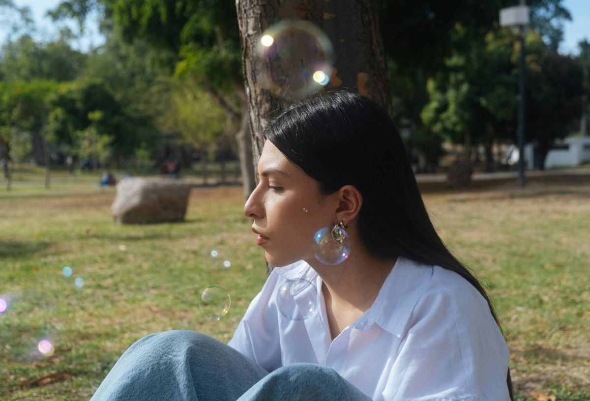 Sötét, hosszú hajú fiatal nő ül fehér ingben és farmerben parkban, körülötte szappanbuborékok