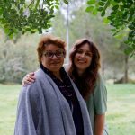 fiatalabb hosszú hajú nő átöleli idősebb szemüveges nő vállát, fa alatt állva a kamerába mosolyognak