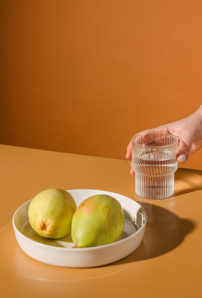 Letisztult fotón barna háttér előtt barna asztalon fehér tálban két körte, egy női kéz nyúl egy vizespohárért