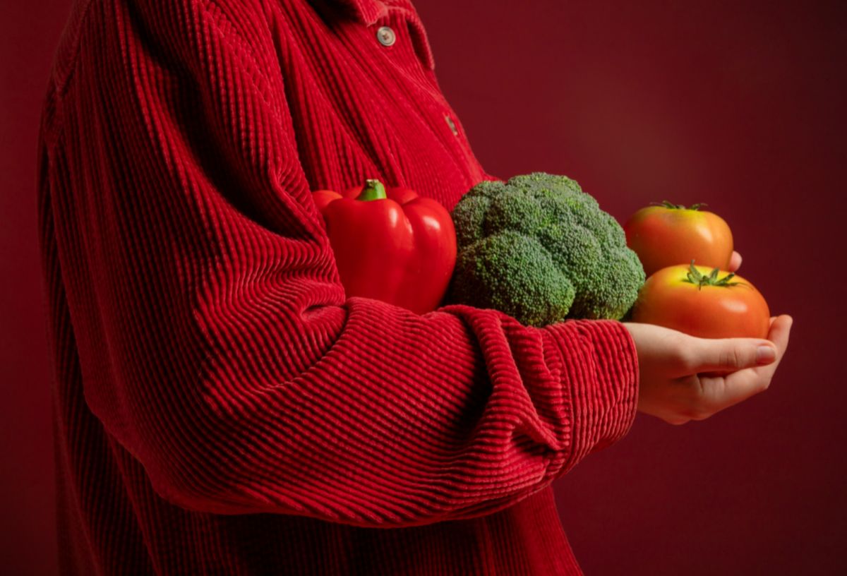 Nő piros kordbársony ingben karjában brokkolit és pritamin paprikát tart