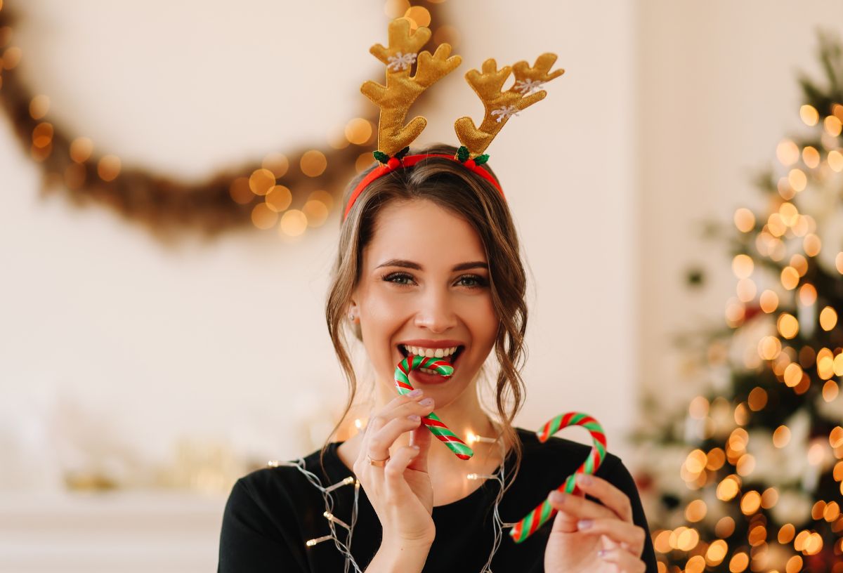 Szép barna hajú nő rénszarvasagancs fejdísszel és fényfüzérrel a nyakában beleharap karácsonyi cukorka pálcába, háttérben elmosódott karácsonyfa