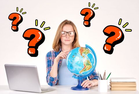 Okos, szőke, fiatal lány flanelingben és fekete keretes szemüvegben íróasztalnál ül, előtte földgömb, ceruázk, laptop, piros kérdőjelek