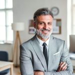 Szürke zakóban egy ősz hajú üzletember mosolyog a kamerába